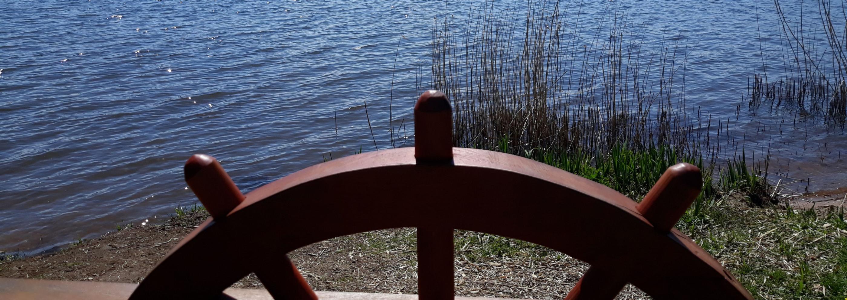 Man sieht ein eingelassenes Schiffssteuer auf einer Balustrade, dahinter der Bostalsee bei Sonnenschein