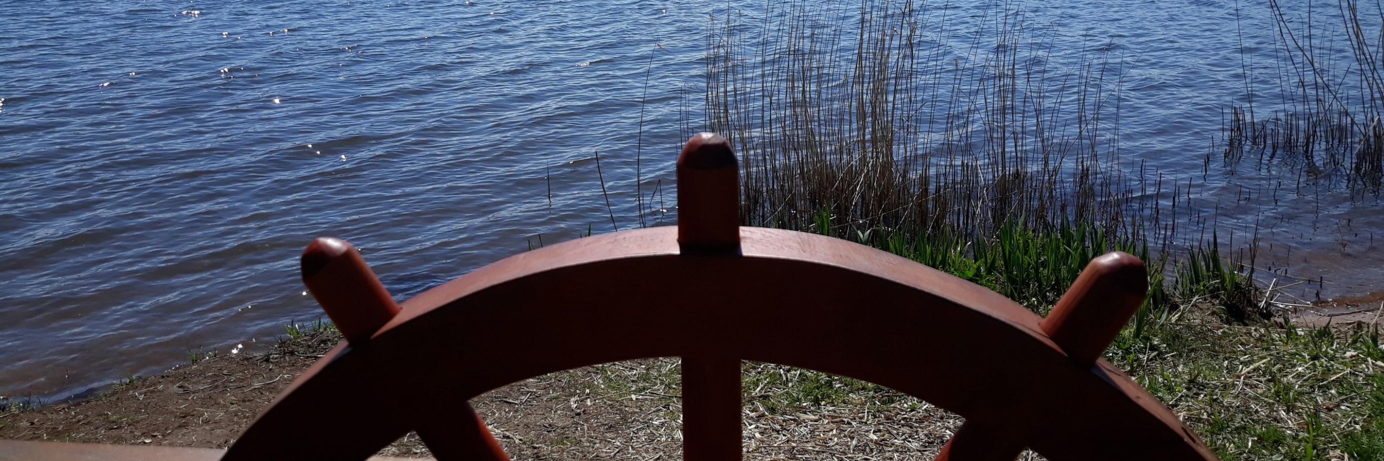 Man sieht ein eingelassenes Schiffssteuer auf einer Balustrade, dahinter der Bostalsee bei Sonnenschein
