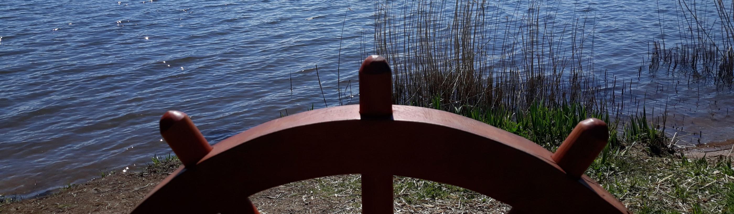Man sieht ein eingelassenes Schiffssteuer auf einer Balustrade, dahinter der Bostalsee bei Sonnenschein